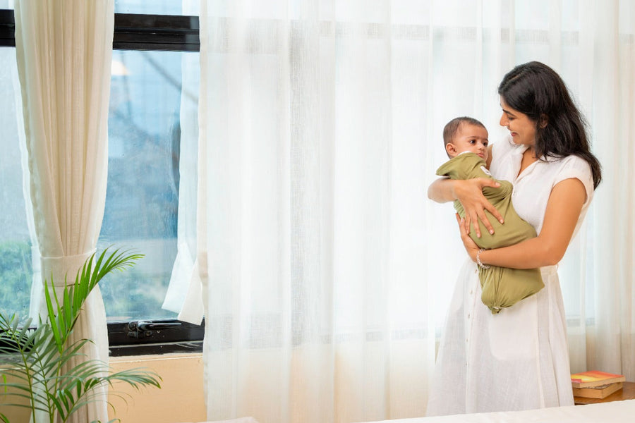 Woman holding a baby in a bedroom with large windows and a plant.