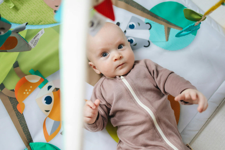 Baby in a brown onesie lying on a colorful play mat with toys.