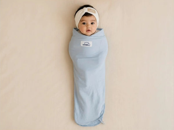 A baby wrapped in a light blue swaddle blanket, lying on a beige surface. The baby is wearing a white headband, and a small white logo patch is visible on the chest of the swaddle.