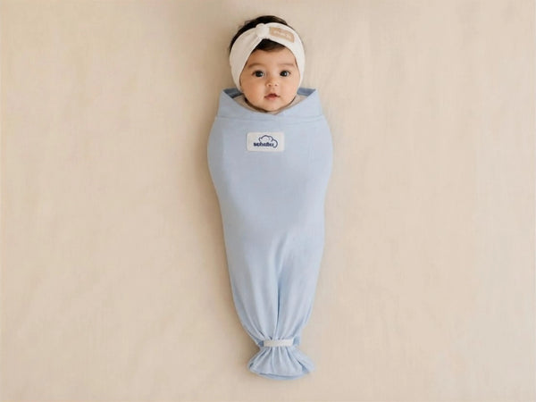 A baby wrapped in a light blue swaddle blanket, lying on a beige fabric background. The baby is wearing a white headband, and a small white brand patch is visible on the chest of the swaddle.