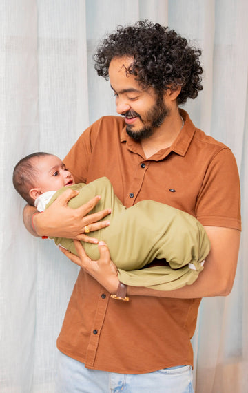 Man holding a baby wrapped in a green blanket against a light curtain background