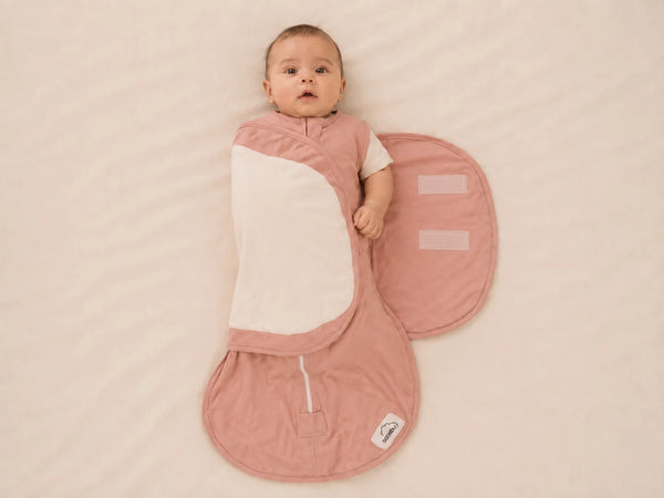 A baby lying on their back wrapped in a dusty pink and white swaddle blanket, placed on a textured cream-colored blanket. The swaddle has a zippered front, a large oval pocket on the side, and a small white brand tag near the bottom.