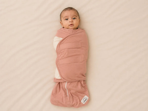 A baby wrapped in a dusty pink swaddle blanket, lying on a textured beige blanket. The swaddle has a white sleeve on one side and a small white brand tag near the bottom.