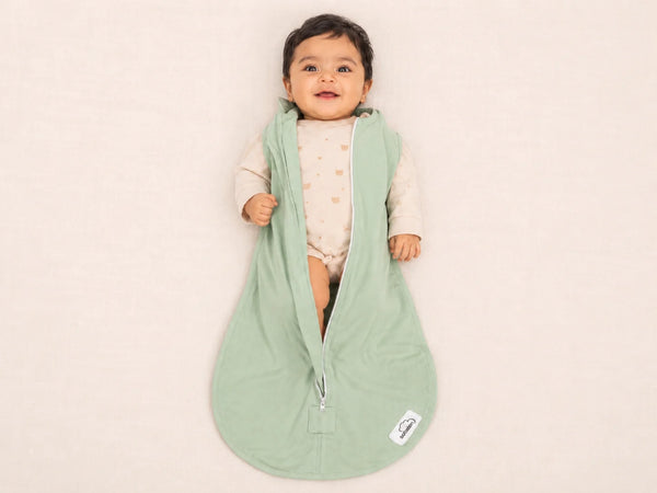 A baby wearing a light sage green zippered sleep sack over a cream bodysuit, lying on a soft cream-colored blanket. The sleep sack has a small white brand tag near the bottom right corner.