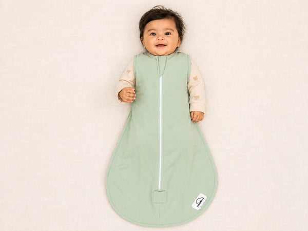 A baby wearing a sage green zippered sleep sack over a light beige long-sleeve shirt, lying on a light beige textured background.