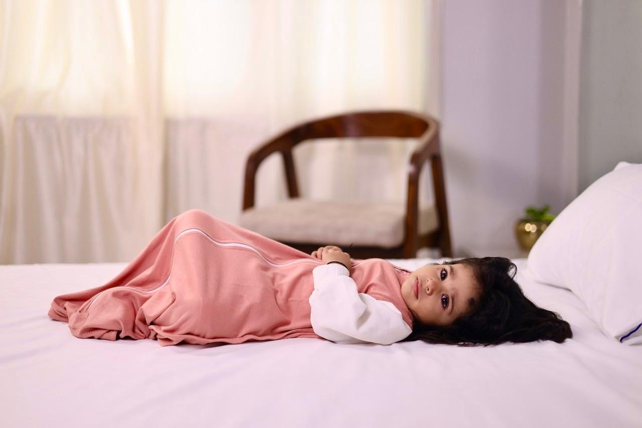 Child in a clay nest peach outfit lying on a bed with a blurred background