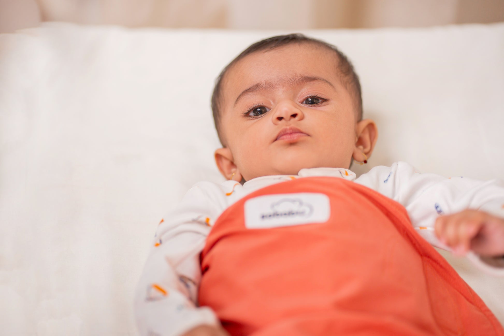 Baby wearing a clay nest peach outfit with a visible brand label, lying on a white surface