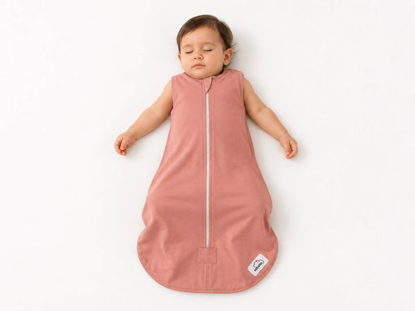 A sleeping baby wearing a dusty rose pink sleeveless sleep sack with a front zipper, lying on a clean white background.