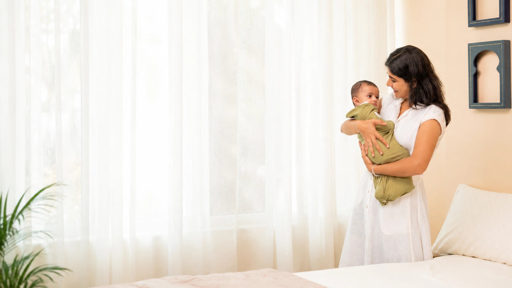 A woman in a white dress holds a baby wrapped in an olive green swaddle blanket in a softly lit bedroom with sheer curtains and a bed.