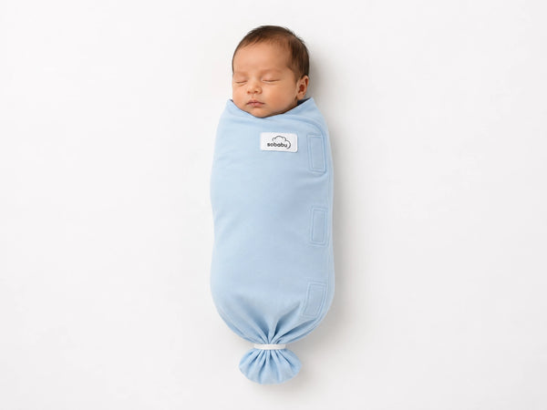 A sleeping newborn baby swaddled in a light blue wrap blanket, shown against a clean white background. The wrap has a small white brand label on the chest and is tied at the bottom.